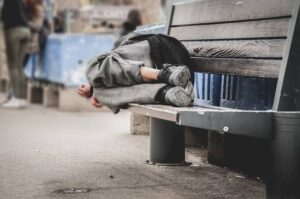 Poor homeless man or refugee sleeping on the wooden bench on the urban street in the city, social documentary concept, selective focus-cm
